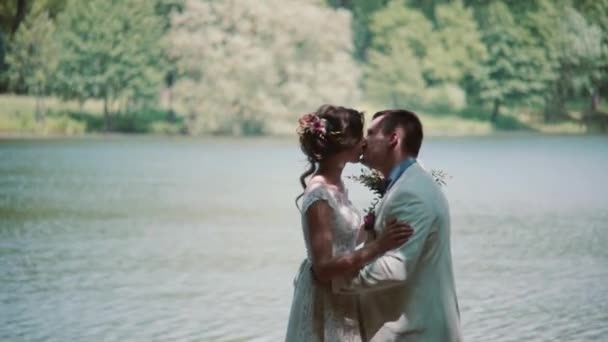 Вид сбоку на целующуюся пару в день их свадьбы. Счастливая невеста и жених смеются в красивом месте на берегу реки . — стоковое видео