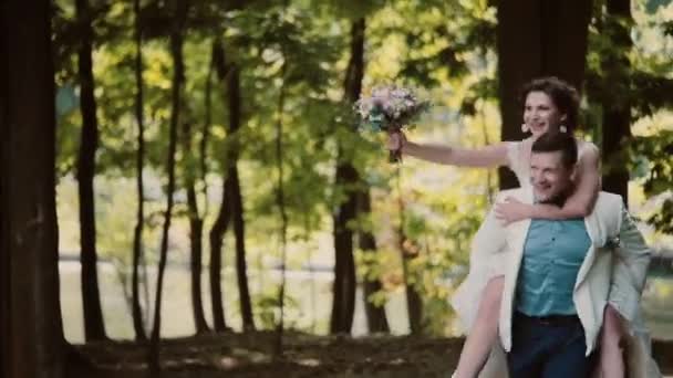Feliz noivo está correndo com sua noiva rindo em suas costas. Casal feliz se divertindo em seu dia do casamento . — Vídeo de Stock
