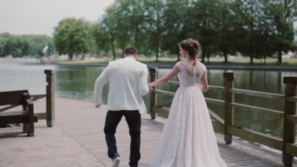 Wunderbares Paar spaziert in Hochzeitskostümen an einem Kai entlang. Sie halten sich an den Händen, unterhalten sich und haben Spaß. schöne Natur. zurück — Stockvideo