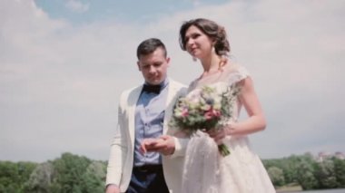 Güzel gelin ve yakışıklı damat yaz aylarında bir parkta yürümek. Beyaz gelinlik ve elbisesi, saç-do. Steadicam atış.