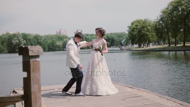 Pareja feliz el día de su boda bailando y divirtiéndose en un muelle. Río en un parque en verano. Trajes de boda — Vídeo de stock