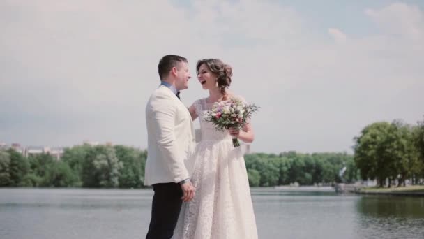 Maravillosa pareja enamorada el día de su boda de pie en una orilla del río, riendo felizmente. Día soleado en verano . — Vídeo de stock