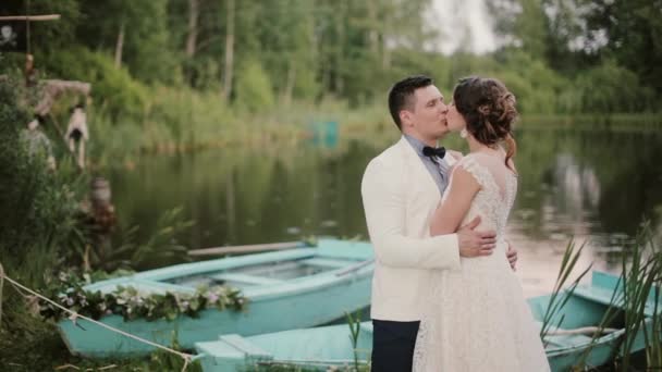 Hermosos amantes se besan cerca de un estanque con barcos azules en él en un lugar hermoso. Novia y novio disfrutan del día de su boda — Vídeo de stock