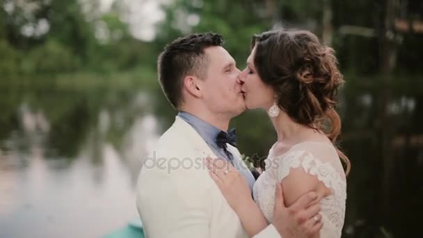 Seitenansicht eines glücklichen Paares, das sich am Hochzeitstag in der Nähe eines Teiches küsst, glücklich lächelt und die gemeinsame Zeit genießt — Stockvideo