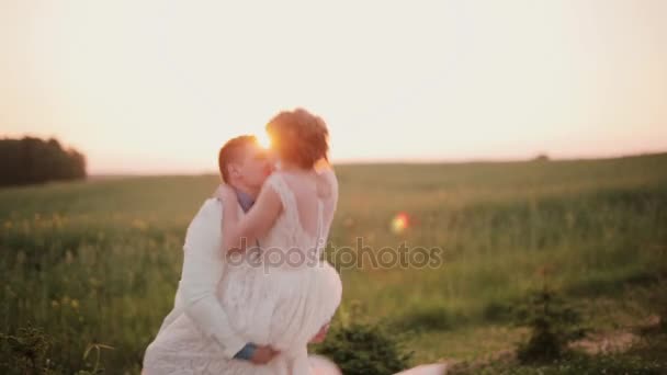 La novia feliz salta a los brazos de sus novios, él la atrapa y gira en un campo al atardecer. Boda en un día de verano . — Vídeo de stock