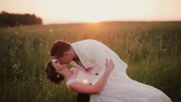 Bonito noivo segura sua bela noiva em seus braços, beijando-a. Ela ri-se. Casamento ao pôr do sol em um lugar bonito — Vídeo de Stock