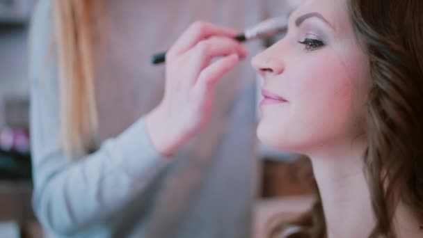 Maquiagem artista aplica-se com escova, use cosméticos decorativos. Visão de close-up da jovem mulher atraente à espera de maquiagem . — Vídeo de Stock