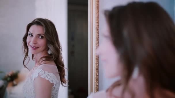 Молода наречена стоїть перед дзеркалом, а потім повертається і дивиться на камеру. Жінка дивиться на відображення перед весіллям . — стокове відео
