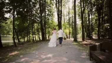 Mutlu bir çift düğün gününde elele bir parkta bir nehir kıyısında yürürken Backview. Güzel beyaz kıyafetler