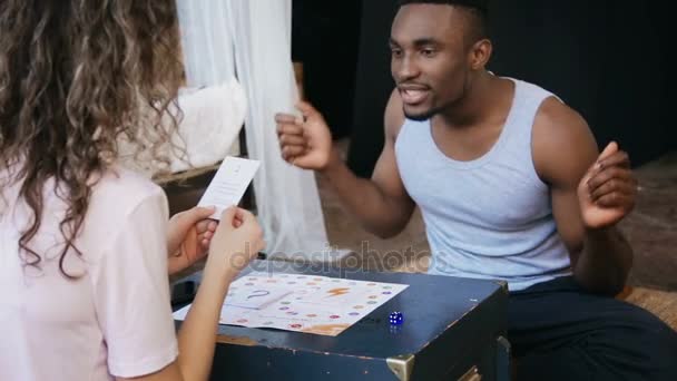 Pareja multiétnica sentada en el suelo y jugando el juego de mesa. Hombre danza mientras que la mujer hace la tarea en tarjeta . — Vídeos de Stock