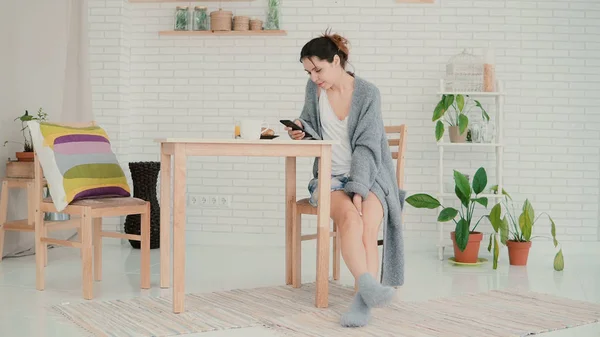 Счастливая женщина сидит на кухне в пижаме и пользуется интернетом. Брюнетка пользуется смартфоном и пьет кофе — стоковое фото