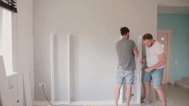 Двое молодых людей делают ремонт в светлой гостиной. Мужчины собирают вместе белую мебель . — стоковое видео