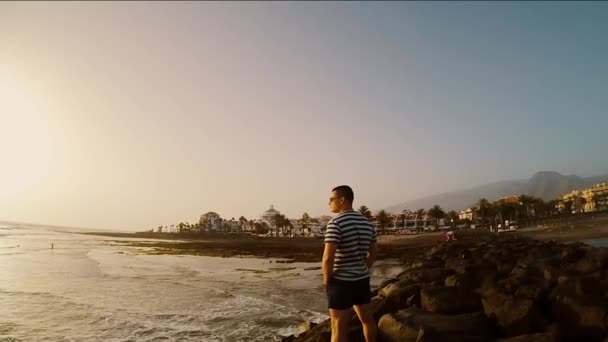 Joven turista de pie en la costa junto al mar en un día ventoso. Hombre mirando a la hermosa puesta de sol . — Vídeos de Stock