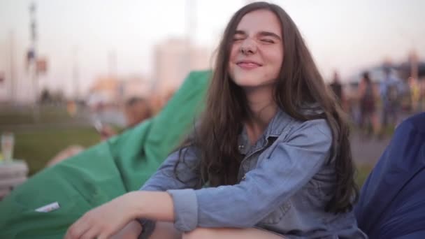 Jonge mooie tiener meisje in jeans jasje glimlachend in het park in warme zomerdag. Zittend in blauwe Boon tas stoel. — Stockvideo