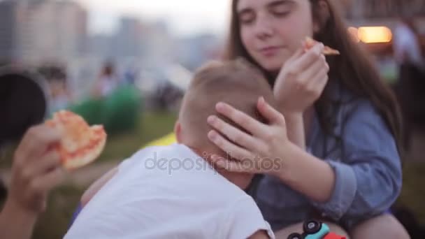 Две девочки и маленький мальчик развлекаются в парке в летний день.Девочки едят пиццу мальчик играет с игрушечным автомобилем . — стоковое видео
