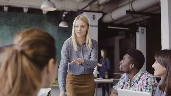 Hermosa mujer rubia gerente dando dirección al equipo multiétnico. Reunión de negocios creativa en la moderna oficina hipster . — Foto de Stock