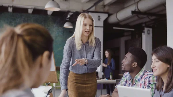 Joven mujer rubia gerente hablando con el equipo de carreras mixtas. Empresaria dando dirección a los colegas en la oficina moderna . — Foto de Stock