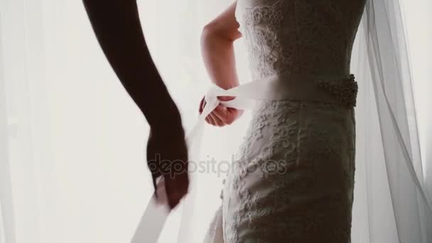 Close-up beeld van jonge bruid zet een boog op haar trouwjurk. Staande in voorzijde van venster en zet op het kostuum vrouw. — Stockvideo