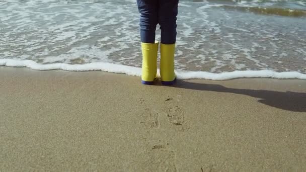 Vista da vicino del piede della bambina in stivali di gomma giallo brillante. Bambino in piedi sulla riva della spiaggia, impronte sulla sabbia . — Video Stock