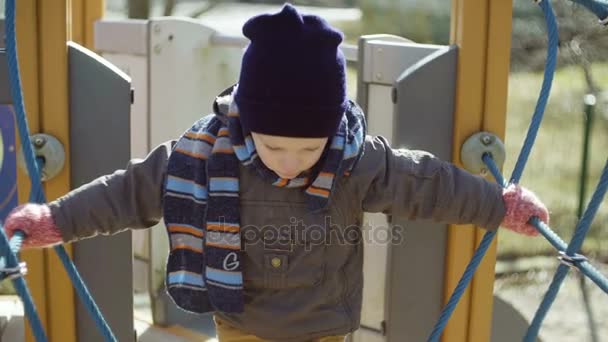 Schattige kleine jongen in oranje rubber laarzen wandelen op speelplaats. Kind maakt zorgvuldig de eerste stappen op kinderen kabelbaan. — Stockvideo