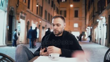 Dışında akşam şehir kafede oturan genç adam. Yakışıklı erkek arkadaşı ile konuşurken kullanmak belgili tanımlık smartphone.
