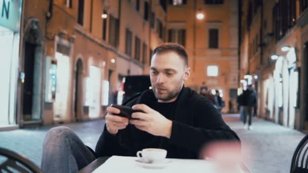 Ung man sitter på kvällen i city café och hålla smartphone. Attraktiv manlig dricka kaffe ensam. — Stockvideo