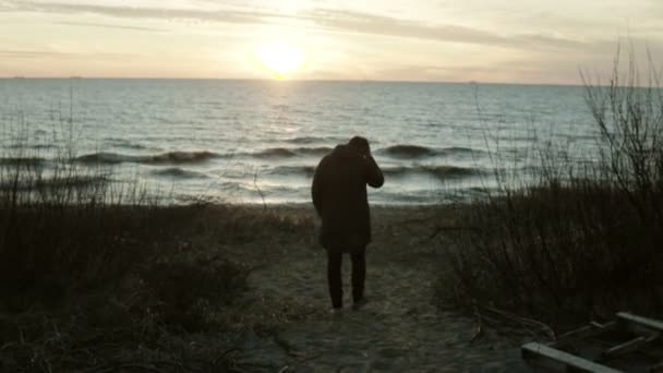 Zadní pohled na mladého muže, chůze na západ slunce na pobřeží moře. Osamělý muž tráví čas o samotě v beach v chladný den. — Stock video