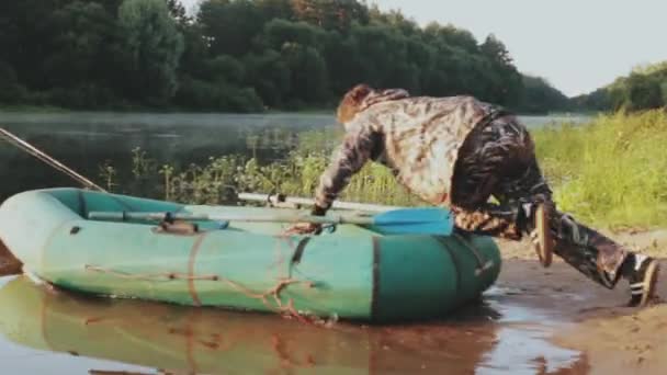 迷彩服の若い男は、水に海岸からゴムボートを置きます。釣りの準備の男性. — ストック動画