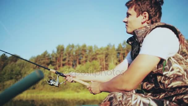 Mladý muž sedí na lodi na jezeře les a vyvolá prut. Atraktivní muž chytání ryb tyčinkou s rotující kotouč. — Stock video