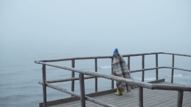 Vista posteriore della bambina in stivali di gomma e plaid in piedi su un molo di legno vicino al mare. Solo bambino nella nebbia giorno ventoso . — Video Stock