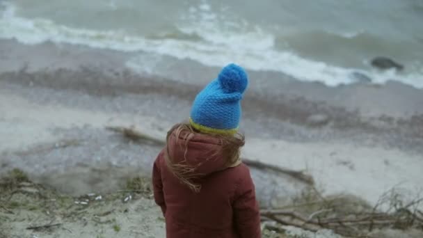 Vista trasera de la niña de pie en la orilla del mar en tiempo frío. Niño solo mirando las olas y soñando — Vídeo de stock