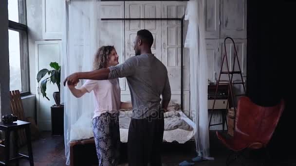 Pareja multiracial en pijama bailando, divirtiéndose juntos por la mañana. La mujer salta sobre las manos del hombre, besos. Movimiento lento . — Vídeos de Stock