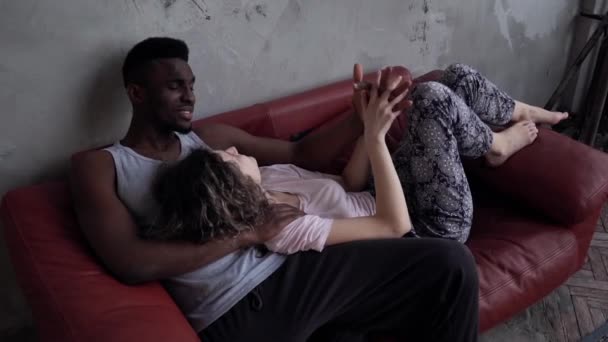 Любляча багатоетнічна пара лежить на червоному дивані і розмовляє. Чоловік і жінка в піжамах тримають руки. Повільний рух . — стокове відео
