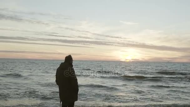 Jovem homem atraente em pé na costa do mar e olhando para o pôr do sol. Masculino apreciando a paisagem na praia . — Vídeo de Stock