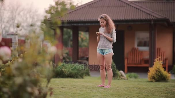 Όμορφη νεαρή γυναίκα στέκεται στο κατώφλι και χρησιμοποιεί το smartphone. Θηλυκό περιηγηθείτε στο Internet από το κινητό. — Αρχείο Βίντεο
