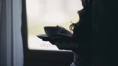 Pencerenin yanında sabahları kahve içme genç çekici kadın silüeti. Kadın mesafe içine görünüyor.