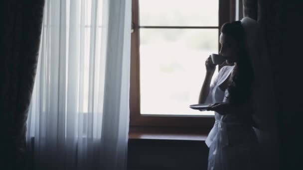 Junge Braut bleibt in der Nähe des Fensters und trinkt Kaffee am Morgen. Frau mit Schleier bereitet sich auf Trauung vor. — Stockvideo