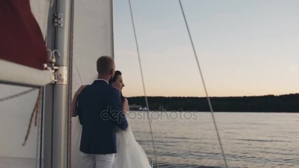 Beautiful newlyweds couple hug, stand on board of sailboat. Bride and groom in wedding outfit looking at distance. — Stock Video