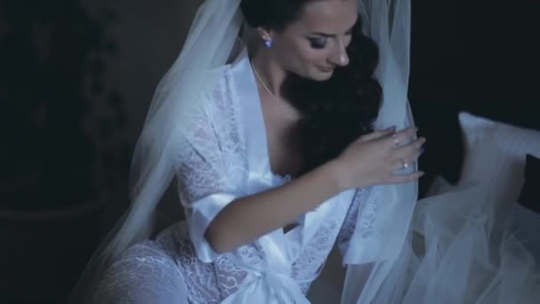 Jeune mariée séduisante en lingerie blanche élégante, peignoir assis sur le lit. Belle femme avec voile posant sur la chambre . — Video