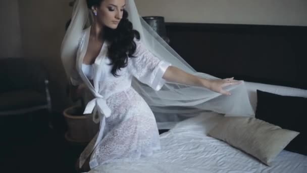 Jeune mariée séduisante en lingerie blanche élégante, peignoir assis sur le lit. Brunette femme fixe son voile et pose .
