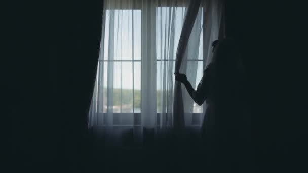 窓の近くに白いペニョワールランジェリーの若い花嫁のシルエット。女性に見える距離で、結婚式前に、の朝. — ストック動画