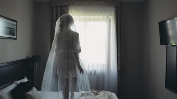 Reggel, a gyönyörű menyasszony. Fiatal nő fehér fehérnemű, peignoir és fátyol tánc az ágyon, esküvő előtt. — Stock videók