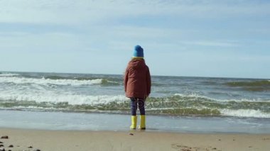 Sevimli küçük kız içinde parlak sarı lastik çizme Beach sahilinde. Çocuk deniz, dalgalardan bakarak onlardan uzak çalıştırmak