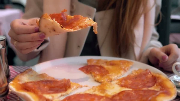 Привлекательная брюнетка держит кусочек пиццы и кусает его. Голодная девушка ужинает в ресторане быстрого питания . — стоковое фото