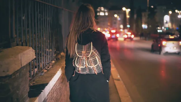 Brunette woman with backpack walking late at night. Attractive girl goes through the city centre near road in evening.