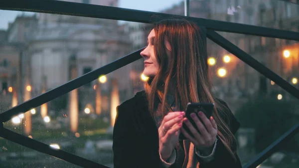 Young beautiful woman sitting in the city at evening and use smartphone. Girl texting with touchscreen keyboard.