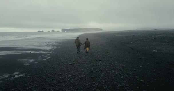 Terug luchtfoto van jonge hipster paar lopen op de zwarte vulkanische strand in IJsland. Man en vrouw genieten van de zee. — Stockvideo