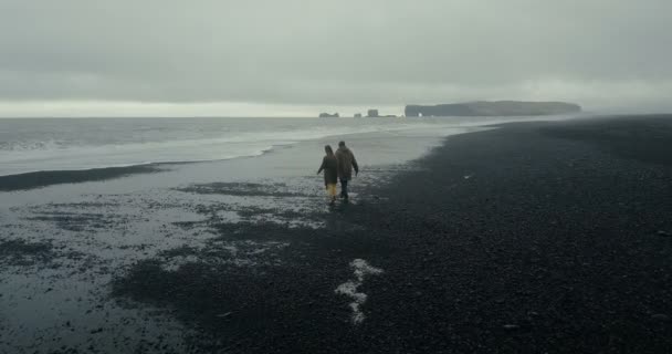 Vue aérienne de jeunes couples élégants marchant sur la plage volcanique noire en Islande. Homme et femme fuyant la vague . — Video