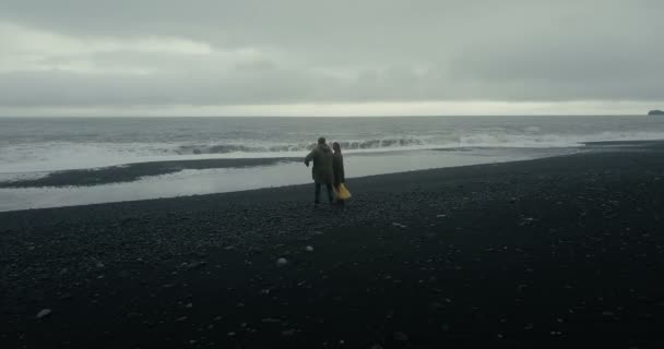 Vista aérea de una joven pareja parada en la playa volcánica negra cerca de las rocas de los dedos de los pies del troll y disfrutando de las olas en Islandia . — Vídeos de Stock