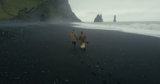 Luchtfoto terug van het echtpaar van de jonge hipster uitgevoerd op de zwarte vulkanische strand in de buurt van de troll tenen in IJsland. — Stockvideo